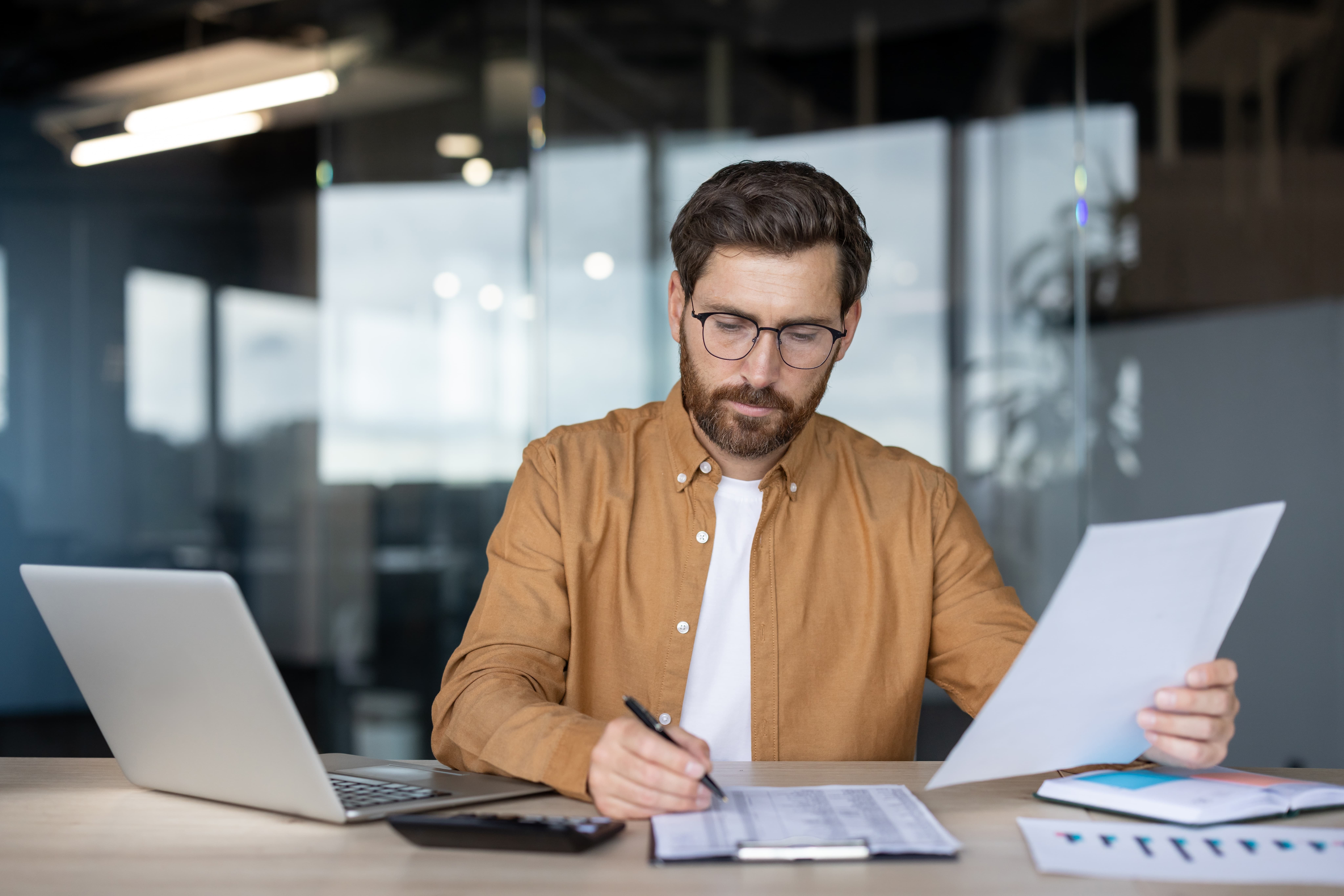 A business man sitting at a desk with an open laptop while doing paper work